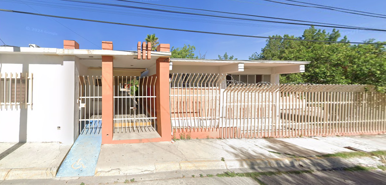 Fotografía en la que se muestra la entrada del plantel, conformada por una reja de color blanco que rodea las instalaciones. Del lado izquierdo se encuentra una rampa y la puerta de acceso.